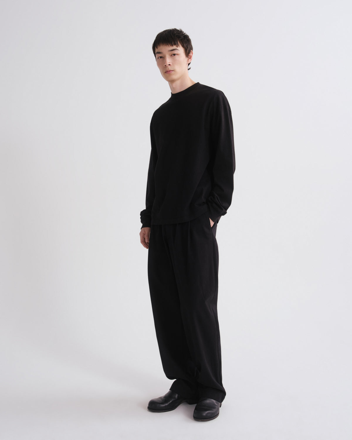 A model wearing a black long-sleeve crewneck top and matching loose black trousers, standing against a plain light gray studio background.
