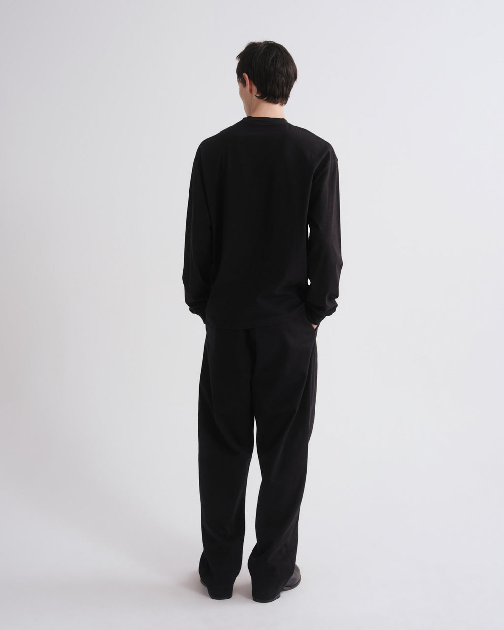 A man viewed from behind wearing a black long-sleeve crewneck top and matching black wide-leg trousers, standing against a plain light gray studio background.