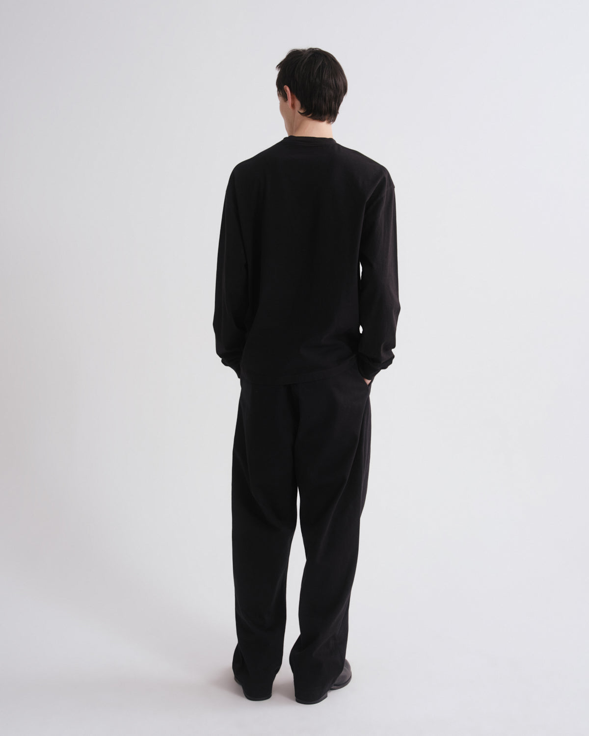 A man viewed from behind wearing a black long-sleeve crewneck top and matching black wide-leg trousers, standing against a plain light gray studio background.