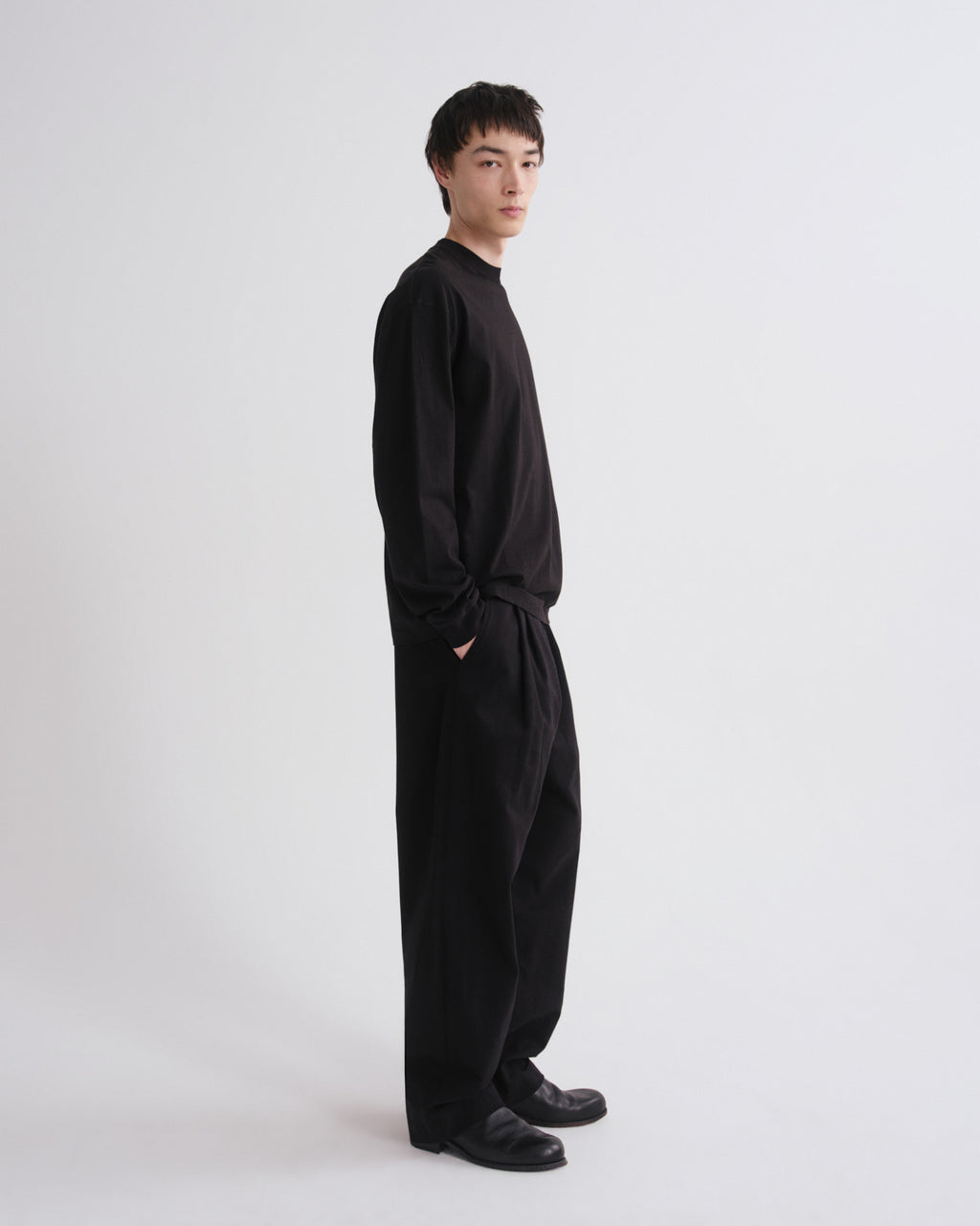 A man wearing a black long-sleeve top and matching black wide-leg pants, standing in profile against a plain light gray studio background.
