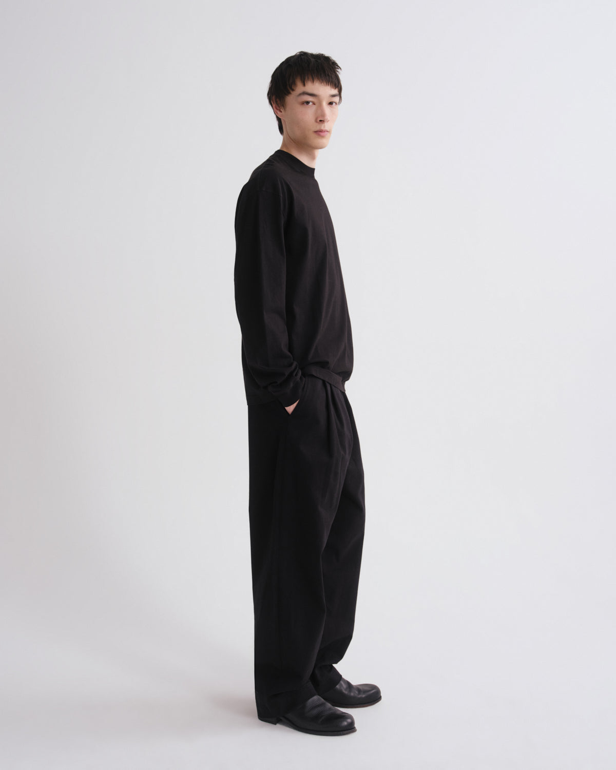 A man wearing a black long-sleeve top and matching black wide-leg pants, standing in profile against a plain light gray studio background.