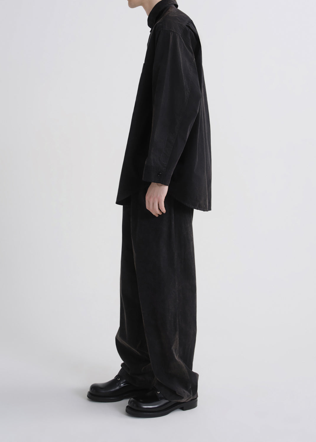 A model shown from the side wearing a black long-sleeve button-up shirt, black wide-leg corduroy trousers, and black leather shoes against a grey background.