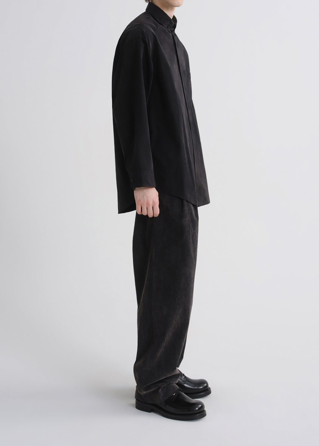 A model shown in profile wearing a black long-sleeve button-up shirt, black wide-leg trousers, and black leather loafers against a grey background.