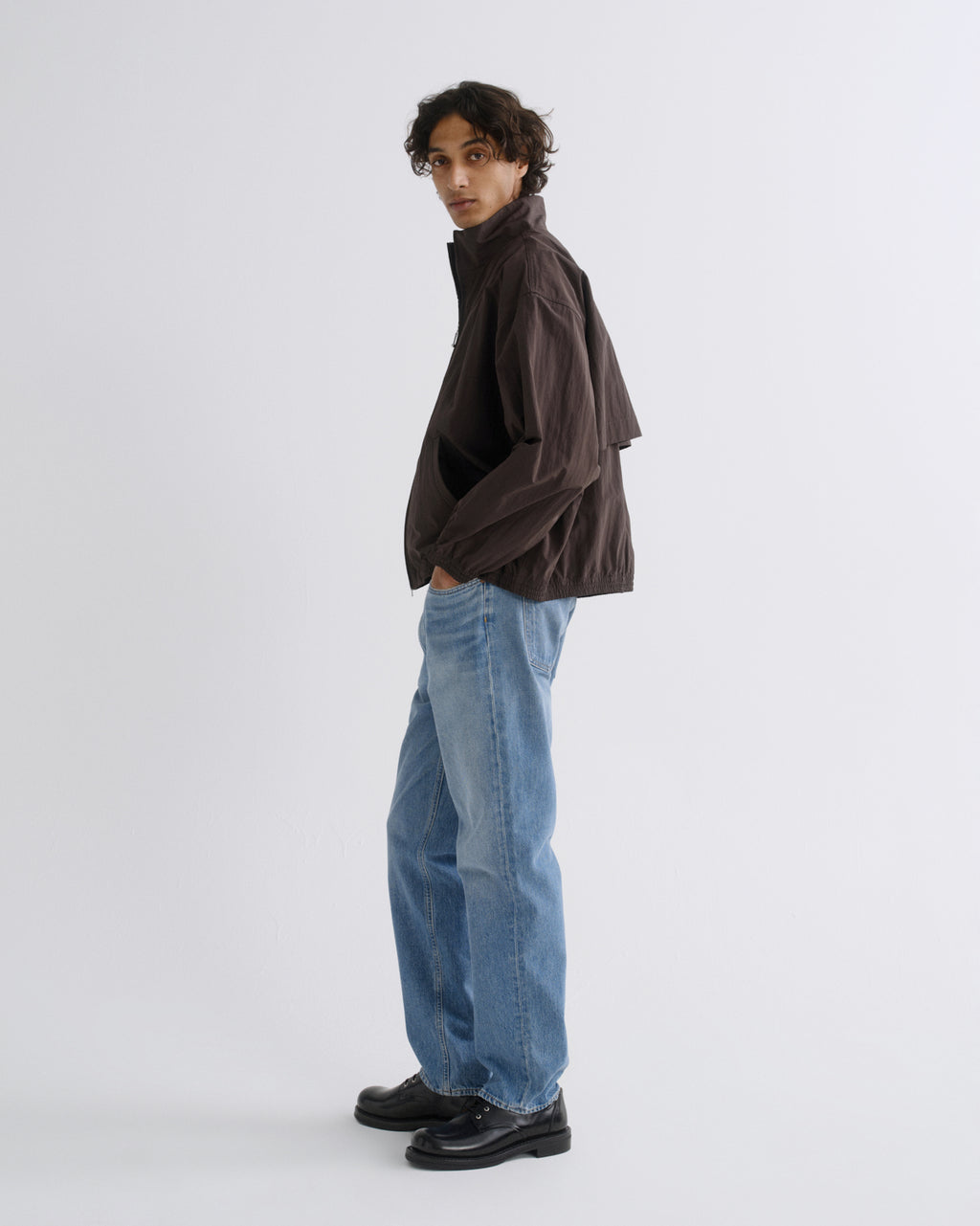 A model wearing a dark brown lightweight jacket, light blue wide-leg jeans, and black leather shoes, standing in profile against a light gray studio background.