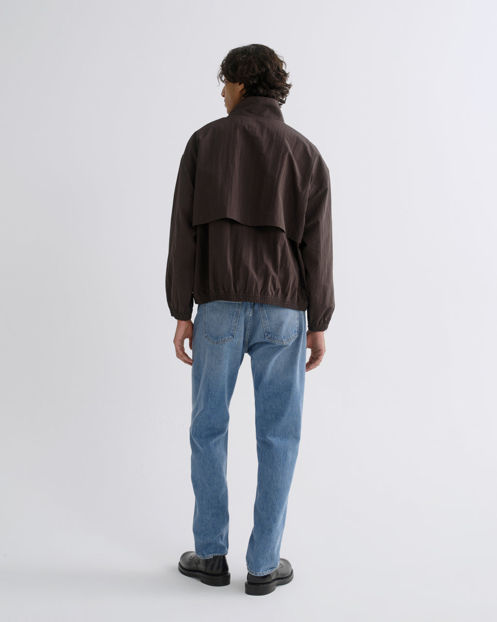A man viewed from behind wearing a dark brown jacket, light blue jeans, and black shoes, standing against a light gray studio background.