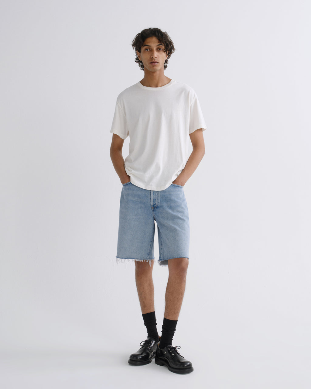 A model stands against a light gray studio background wearing a white crew-neck t-shirt, light-wash denim shorts with raw hems, black socks, and black leather shoes.