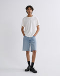 A model stands against a light gray studio background wearing a white crew-neck t-shirt, light-wash denim shorts with raw hems, black socks, and black leather shoes.