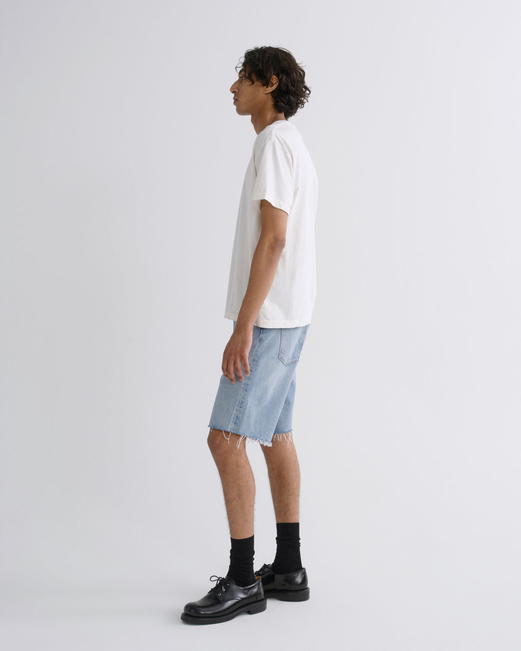 A man shown in profile wearing a white short-sleeve t-shirt, light-wash denim shorts, black socks, and black leather shoes against a plain light gray studio background.