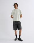 A man modeling a light sage green short-sleeve t-shirt, dark gray denim shorts, black socks, and black leather shoes against a plain light gray studio background.