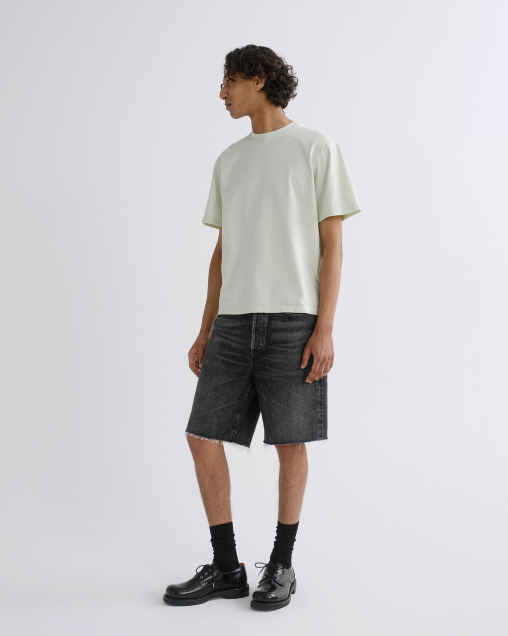 A model stands in profile wearing a pale mint green short-sleeve t-shirt, dark gray washed denim shorts, black socks, and black leather shoes against a light gray studio background.