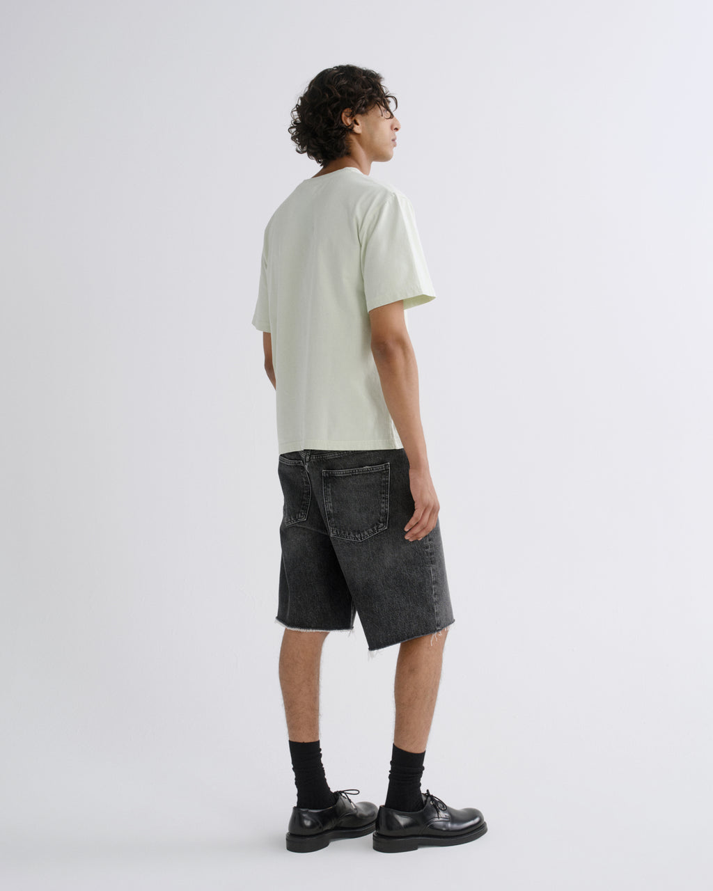 A model shown from the back wearing a pale sage green t-shirt, dark gray washed denim shorts, black crew socks, and black leather shoes, standing against a light gray studio background.