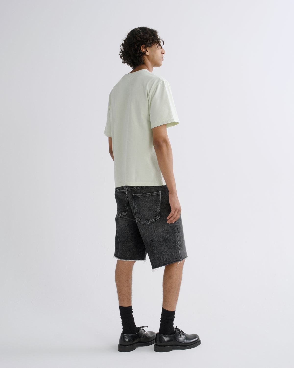 A model shown from the back wearing a pale sage green t-shirt, dark gray washed denim shorts, black crew socks, and black leather shoes, standing against a light gray studio background.