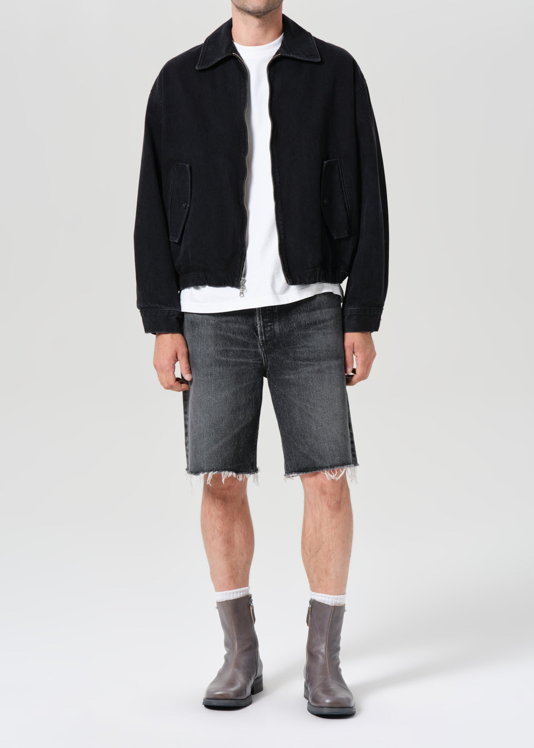 Person wearing a black jacket, white shirt, gray shorts, and gray boots on a white background