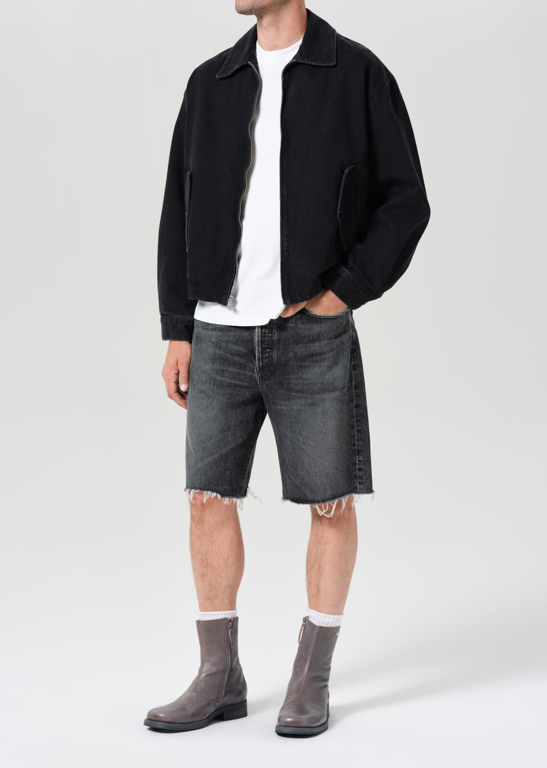 Person wearing a black jacket, white shirt, gray shorts, and gray boots on a white background