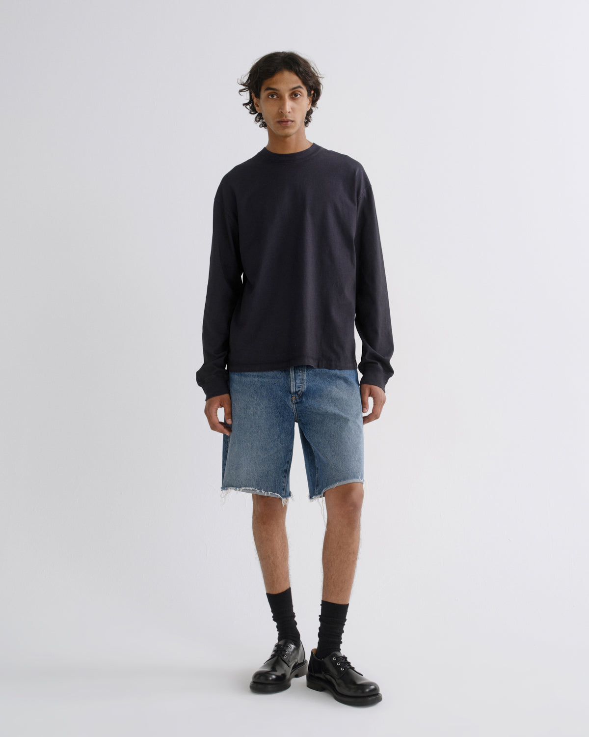 A model stands against a light gray studio background wearing a dark navy long-sleeve crewneck sweatshirt, light-wash blue denim shorts with a raw hem, black socks, and black leather shoes.