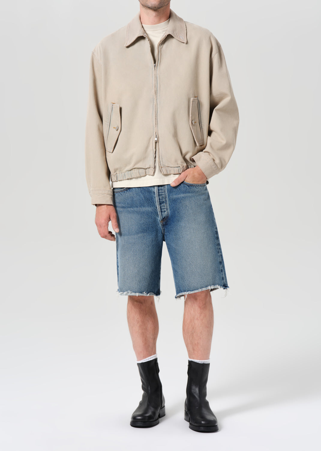 Person wearing a beige jacket, white shirt, blue denim shorts, and black boots on a white background
