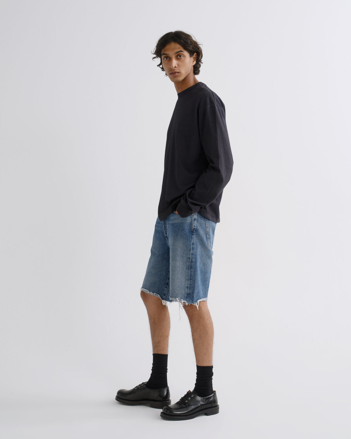 A model wearing a dark navy long-sleeve shirt, light-wash denim shorts, black socks, and black leather shoes, standing against a light gray studio background.
