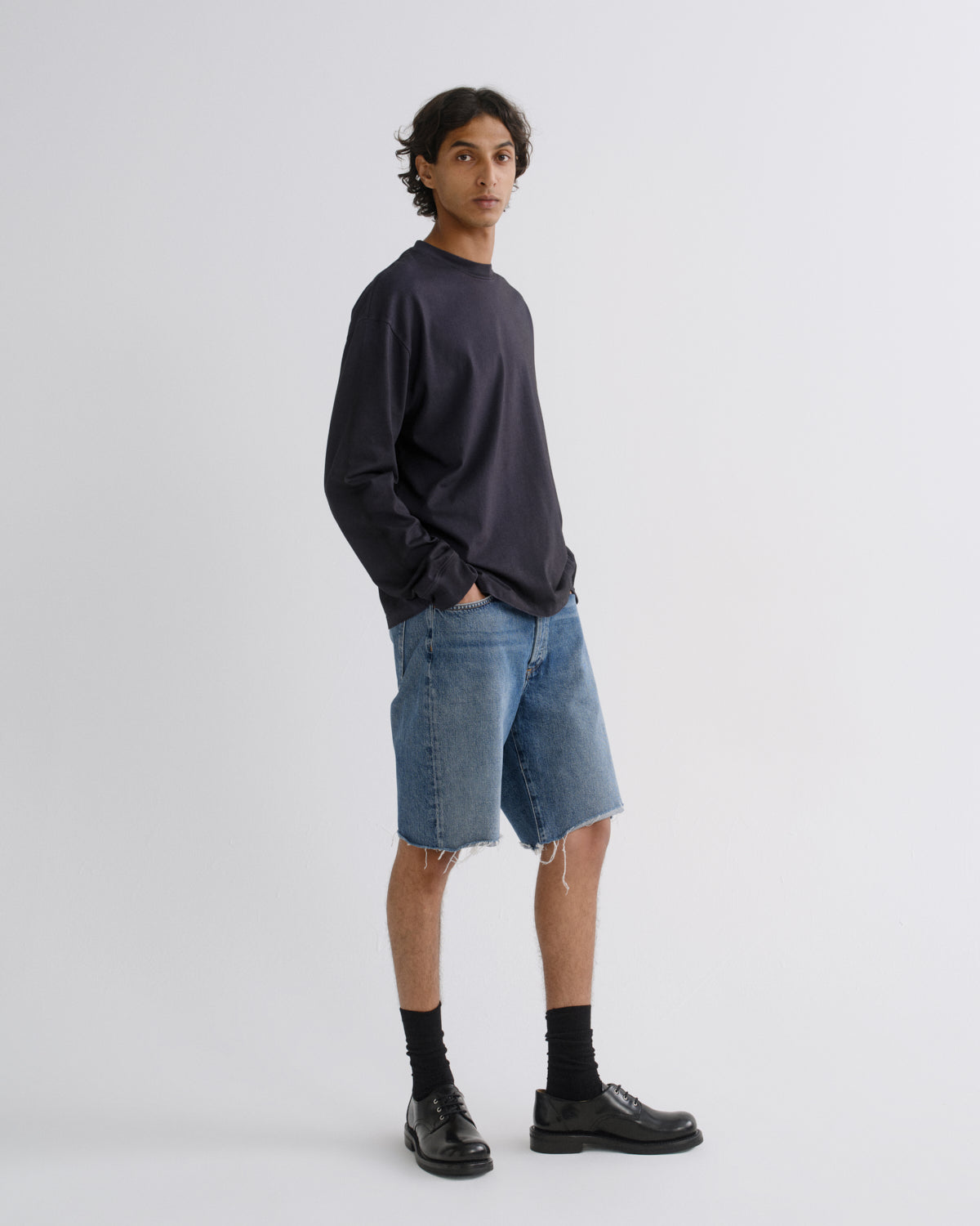 A man modeling a dark navy long-sleeve crewneck sweatshirt, light-wash blue denim shorts with raw frayed hems, black crew socks, and black leather shoes against a light gray studio background.