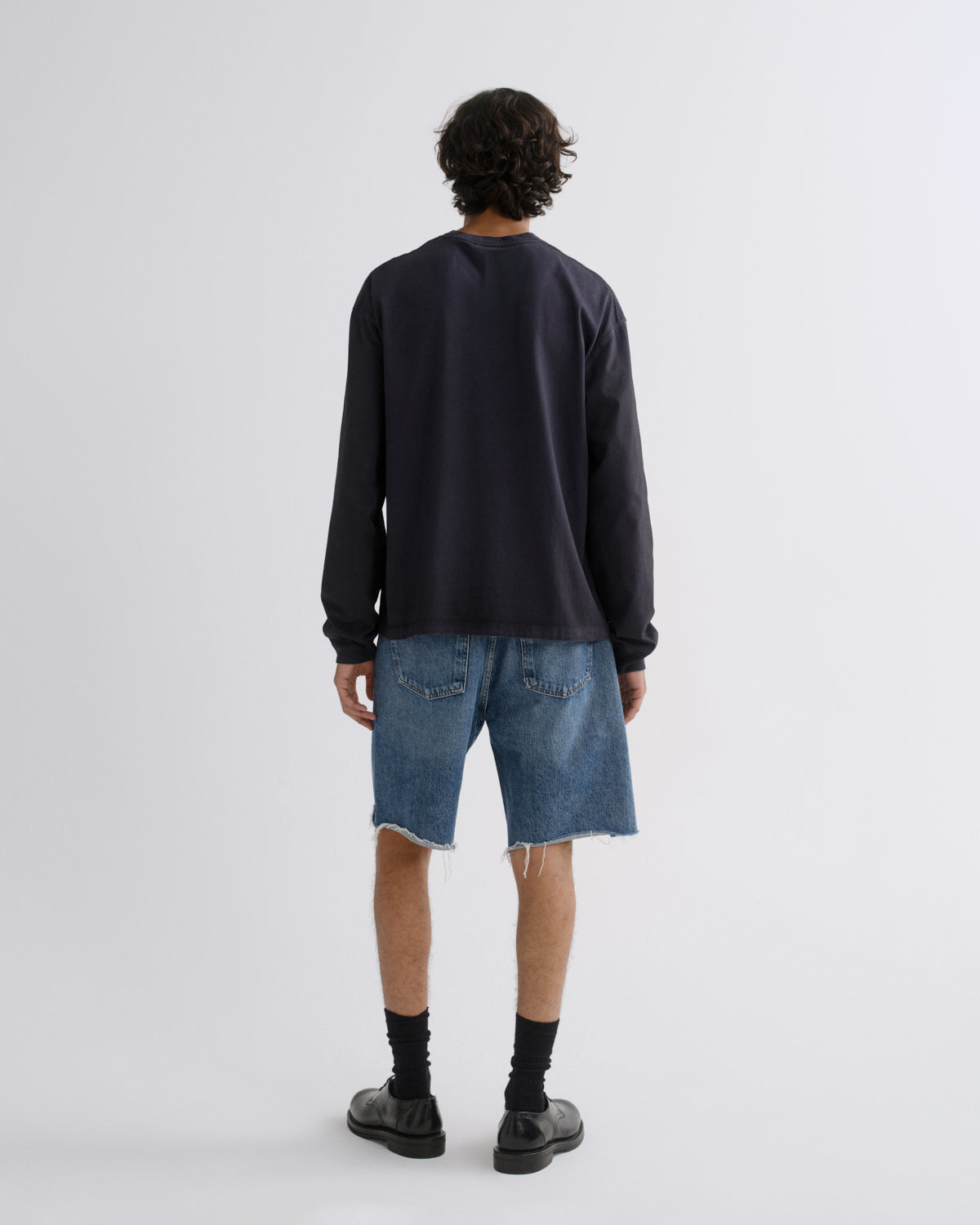 A model shown from the back wearing a dark navy long-sleeve crewneck sweatshirt, blue denim shorts with raw hems, black socks, and black leather shoes, against a light gray studio background.