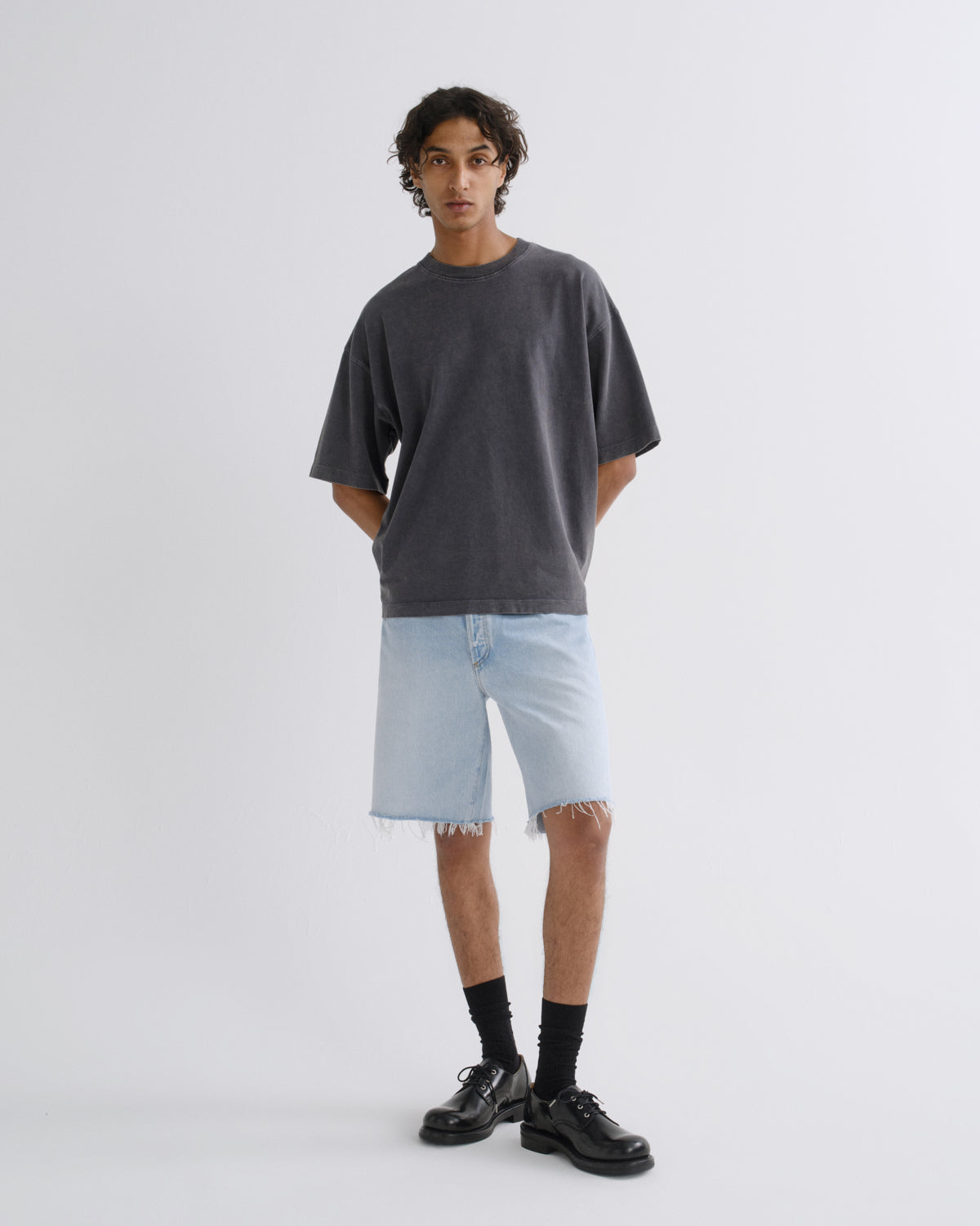 A model stands against a light gray studio background wearing an oversized dark charcoal gray t-shirt, light-wash denim shorts with raw frayed hems, black crew socks, and black leather shoes.