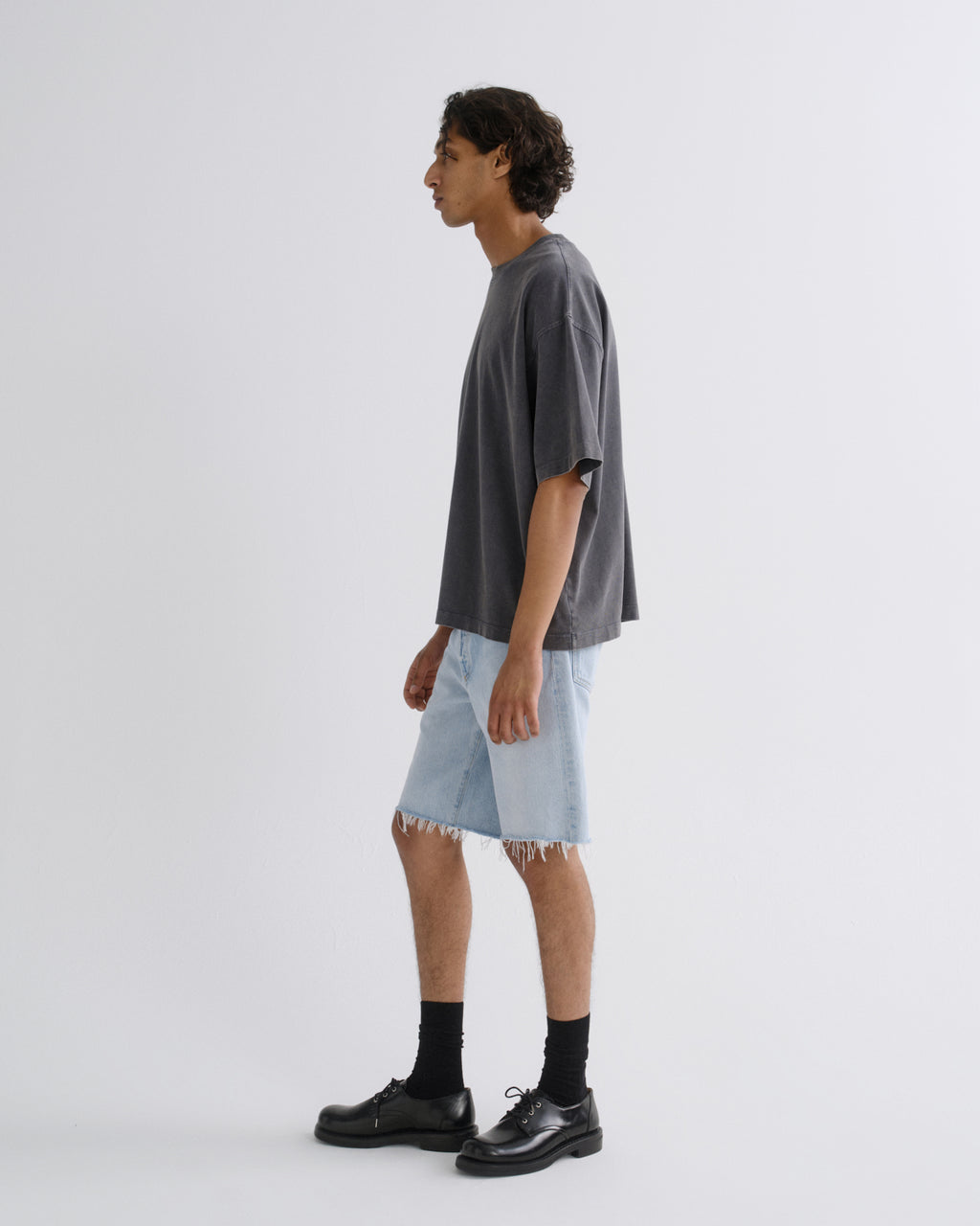 A man in profile wearing an oversized dark gray t-shirt, light-wash frayed denim shorts, black socks, and black leather shoes, standing against a plain light gray studio background.