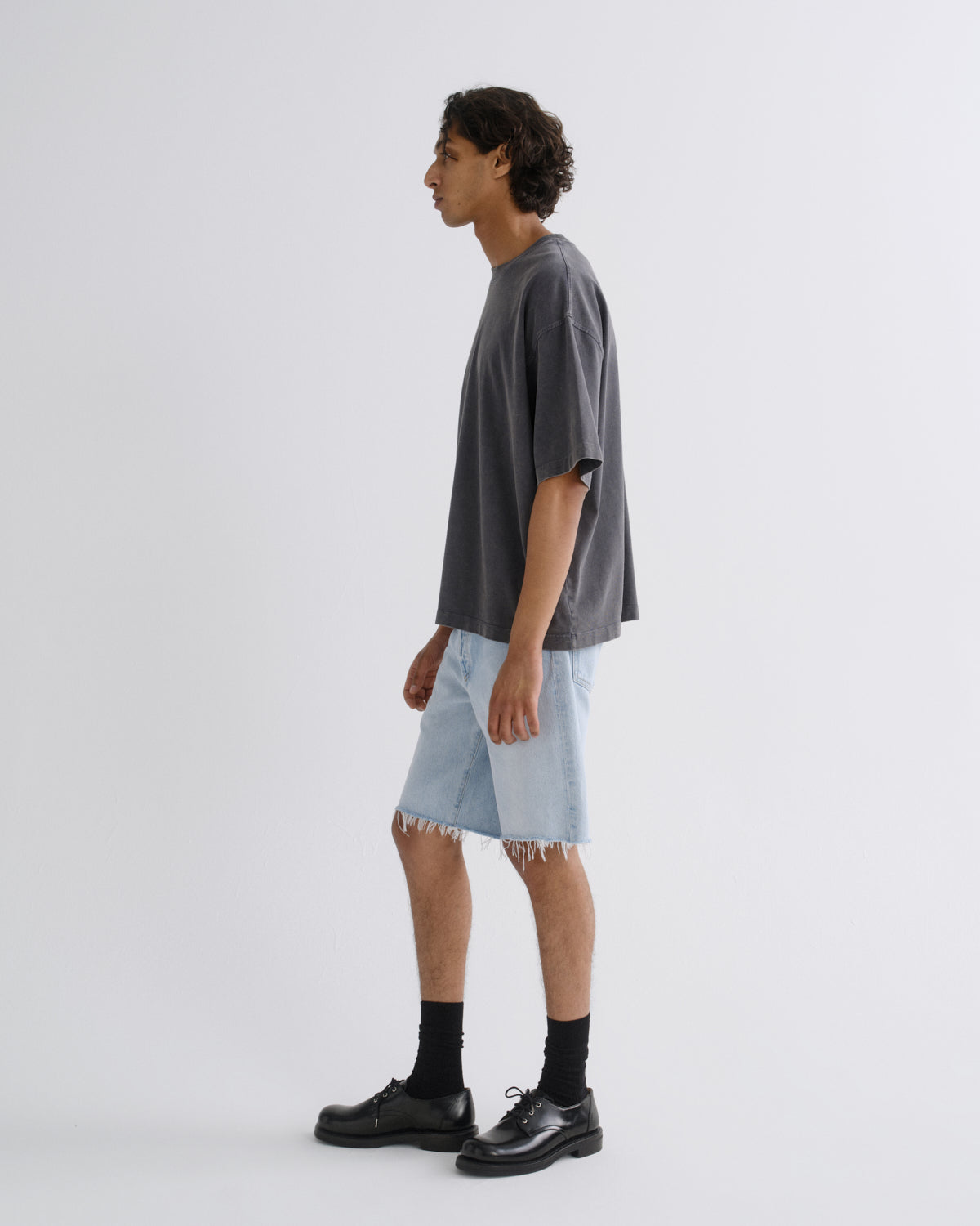 A man in profile wearing an oversized dark gray t-shirt, light-wash frayed denim shorts, black socks, and black leather shoes, standing against a plain light gray studio background.