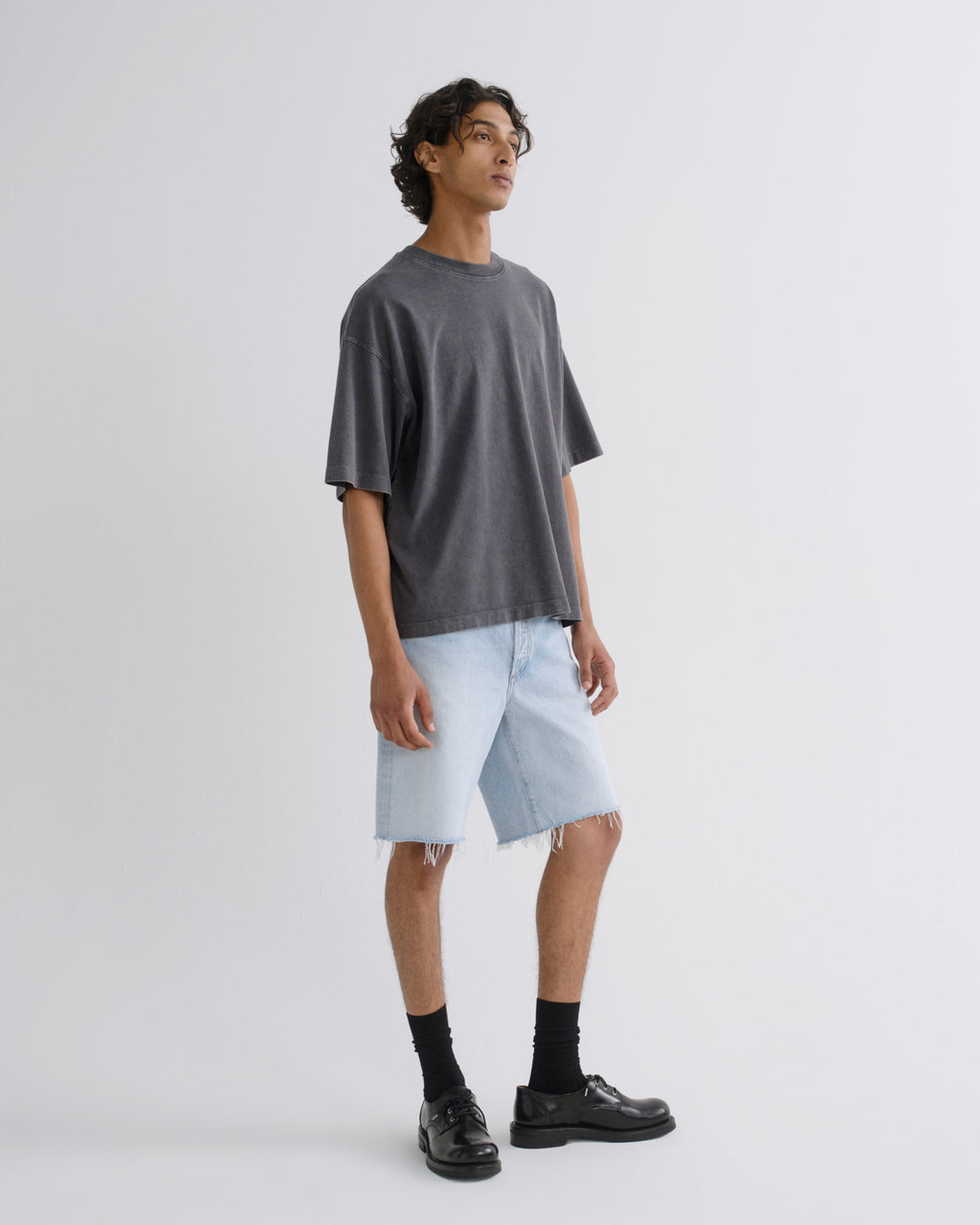 A man modeling an oversized dark gray t-shirt, light-wash denim shorts, black socks, and black leather shoes against a plain light gray studio background.