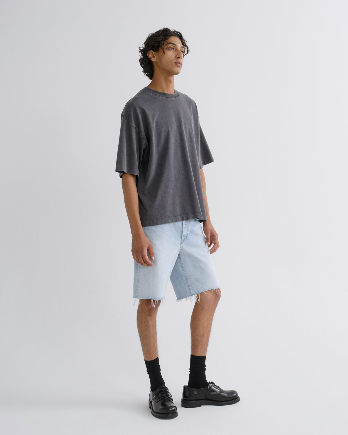 A man modeling an oversized dark gray t-shirt, light-wash denim shorts, black socks, and black leather shoes against a plain light gray studio background.