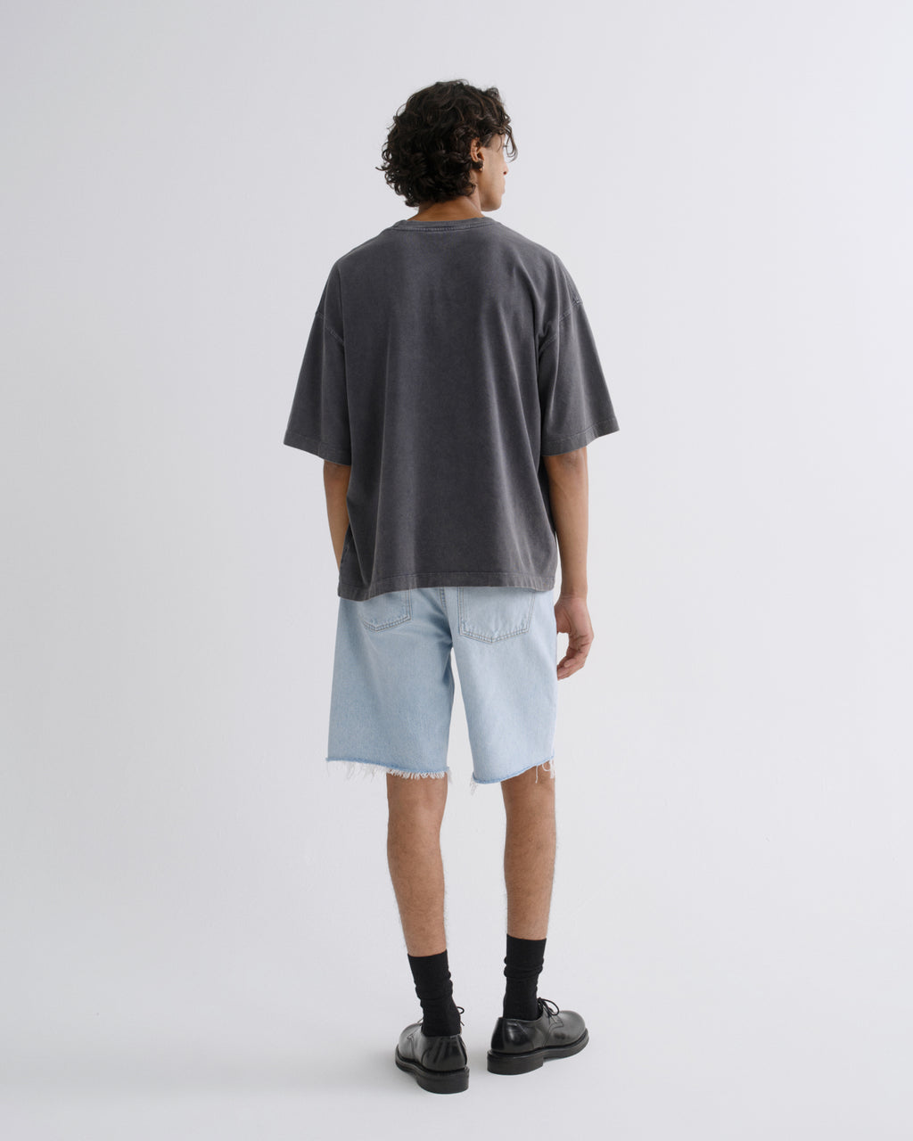 A person shown from the back wearing an oversized dark charcoal gray t-shirt, light wash denim shorts, black socks, and black leather shoes, standing against a light gray studio background.