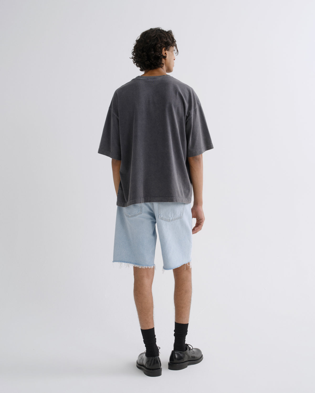 A person shown from the back wearing an oversized dark charcoal gray t-shirt, light wash denim shorts, black socks, and black leather shoes, standing against a light gray studio background.