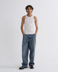 A model stands against a light gray studio background wearing a white ribbed tank top, loose blue jeans, and black loafers.