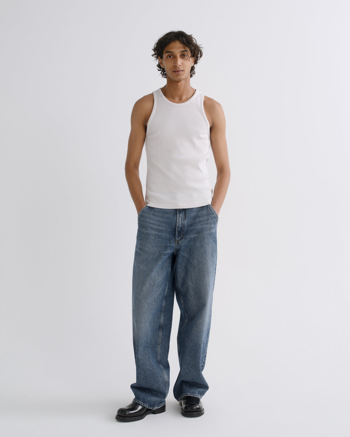 A model stands against a light gray studio background wearing a white ribbed tank top, loose blue jeans, and black loafers.