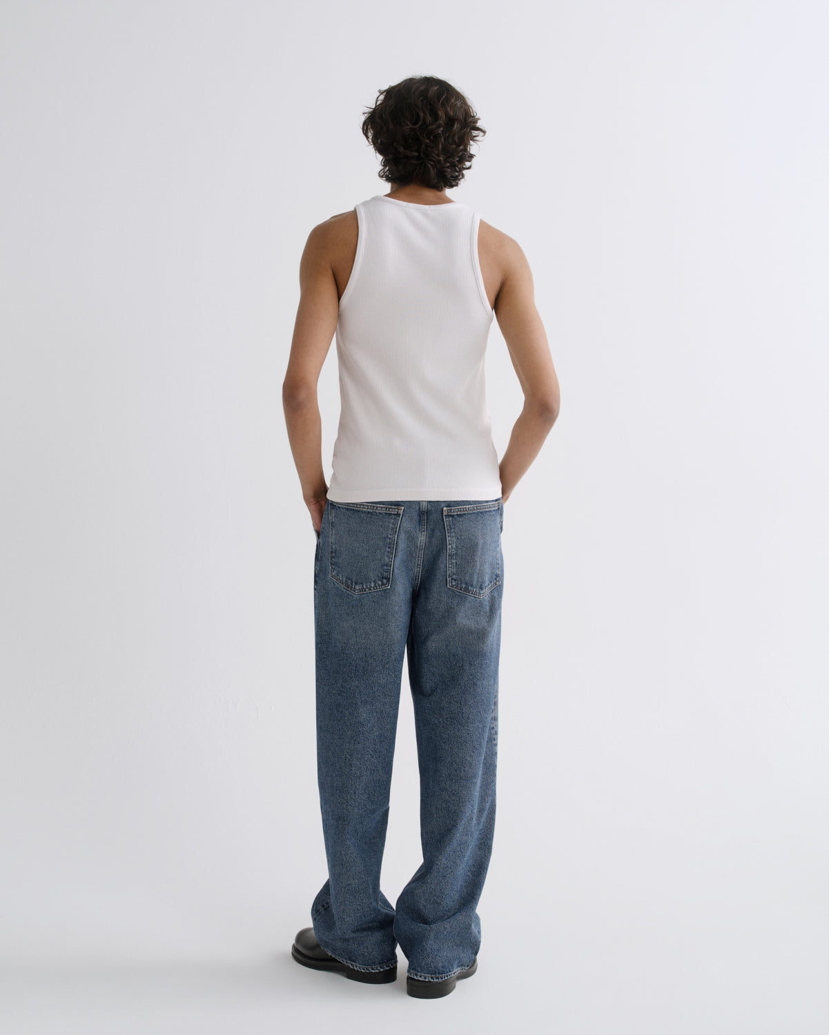 A model shown from the back wearing a white ribbed tank top and wide-leg blue jeans against a plain light gray studio background.