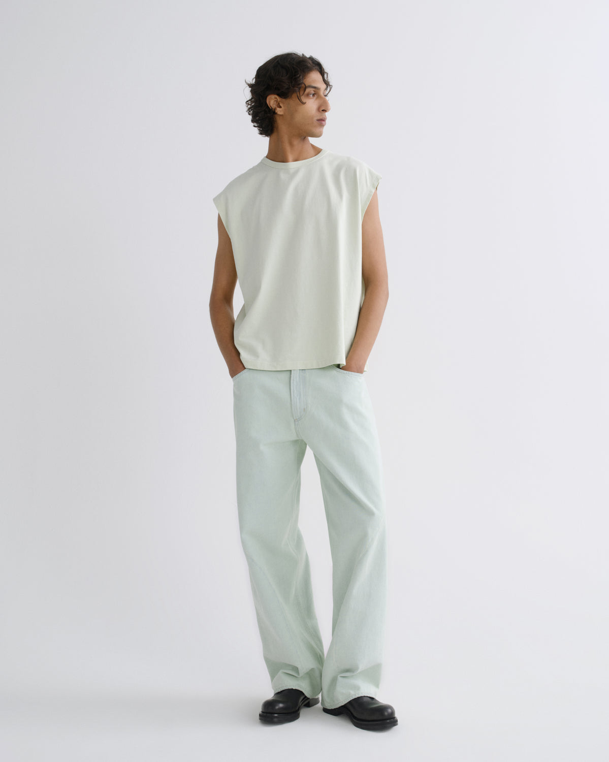 A man wearing a pale sage green sleeveless t-shirt and matching wide-leg trousers, standing against a light gray studio background.