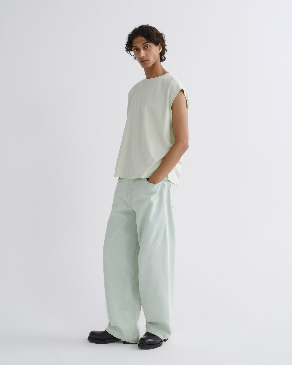 A model wearing a light sage green sleeveless top and matching wide-leg trousers, paired with black shoes, standing against a plain light gray studio background.