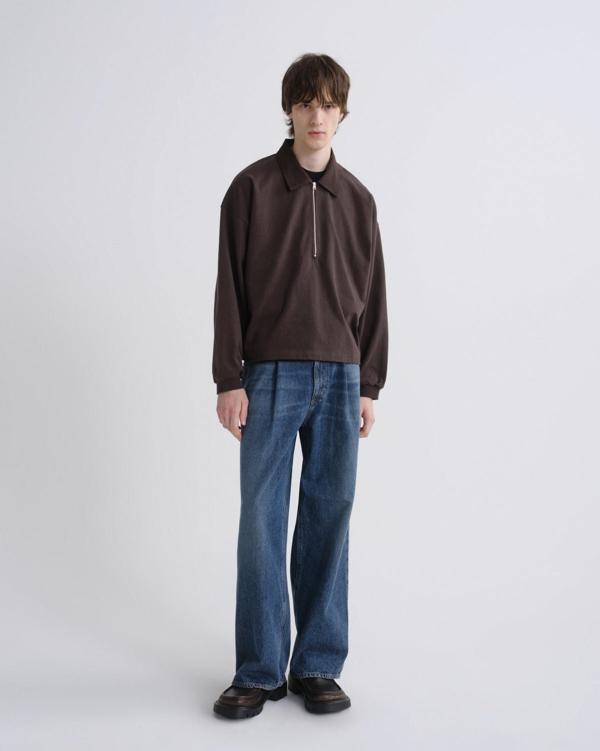 A model stands against a light gray studio background wearing a dark brown long-sleeve half-zip pullover, blue wide-leg jeans, and black shoes.