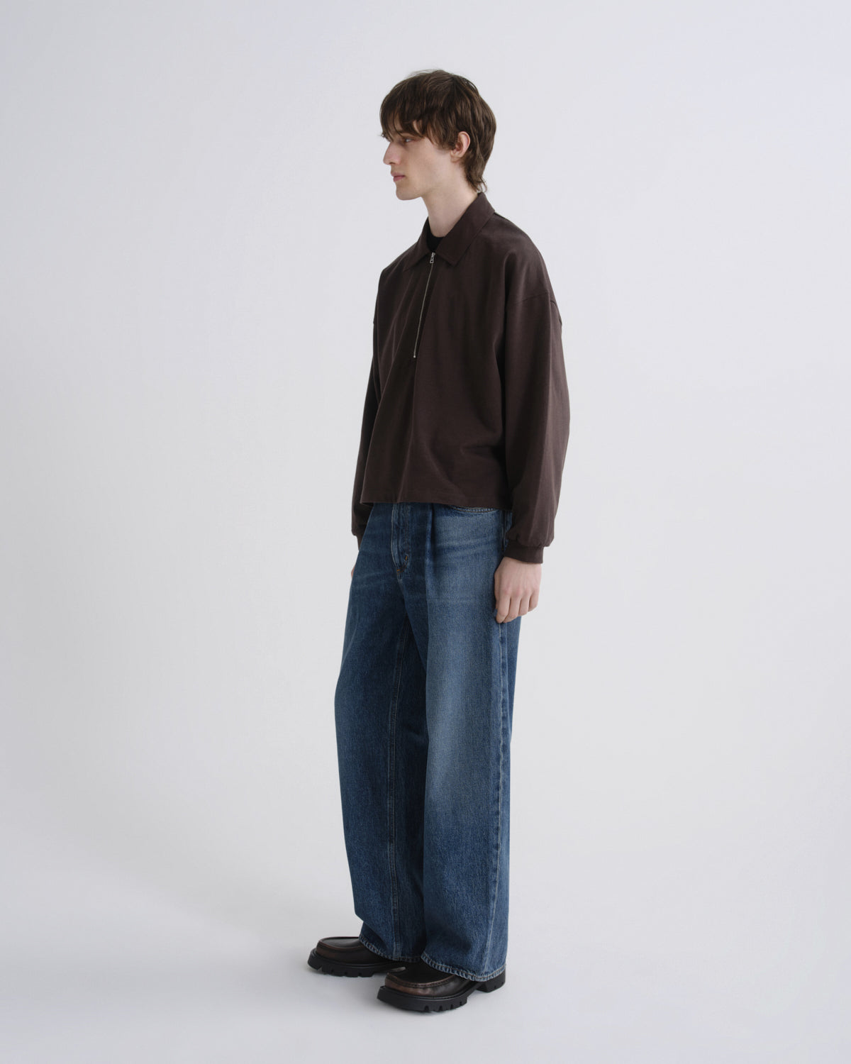 A model stands in profile wearing a dark brown mock-neck zip-up sweatshirt, wide-leg blue jeans, and dark brown shoes against a plain light gray studio background.