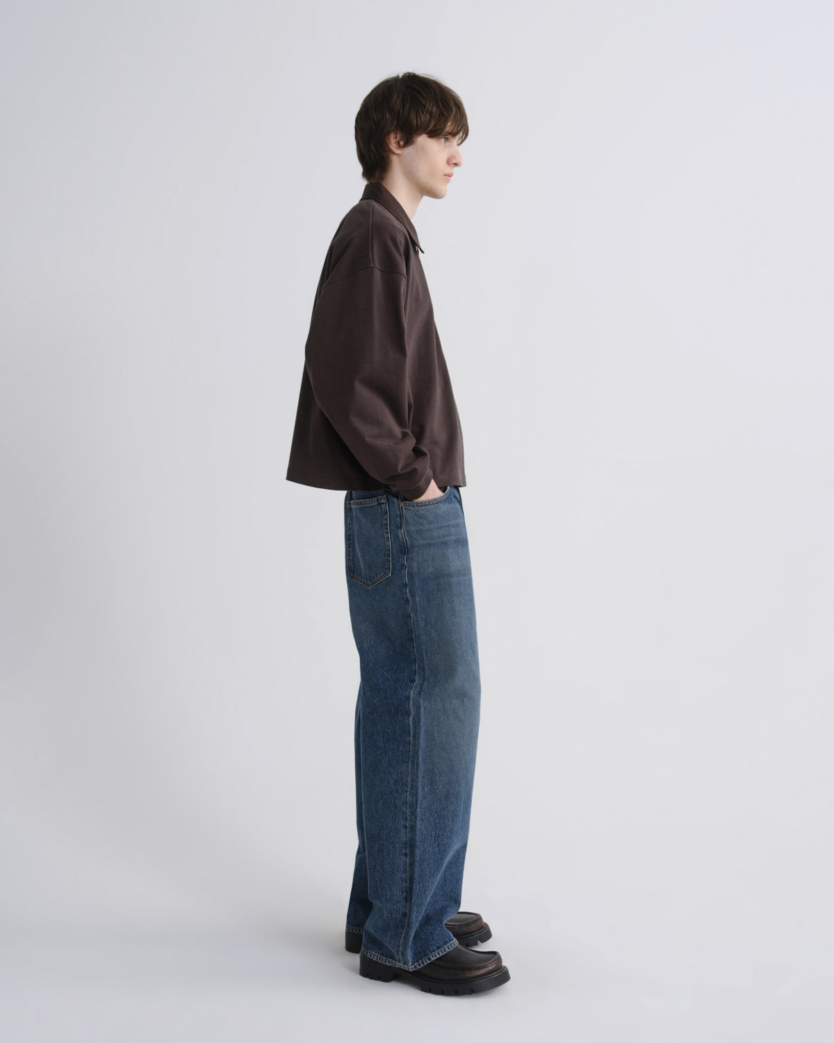 A model standing in profile wearing a dark brown button-up shirt, blue wide-leg jeans, and dark brown loafers against a light gray studio background.