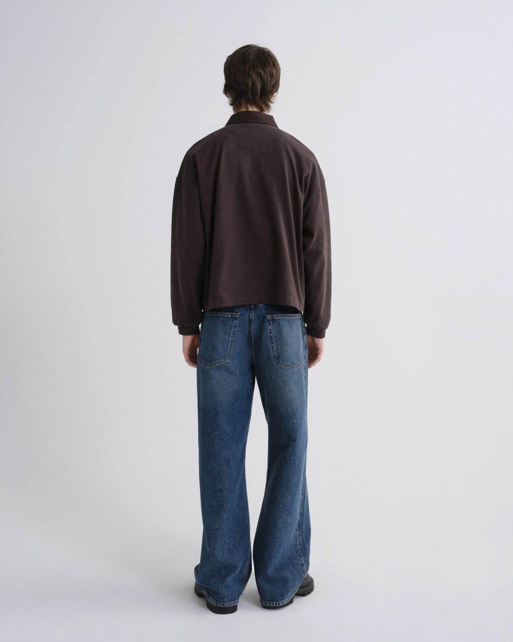 A model shown from the back wearing a dark brown long-sleeve quarter-zip fleece and blue wide-leg jeans, standing against a light gray studio background.