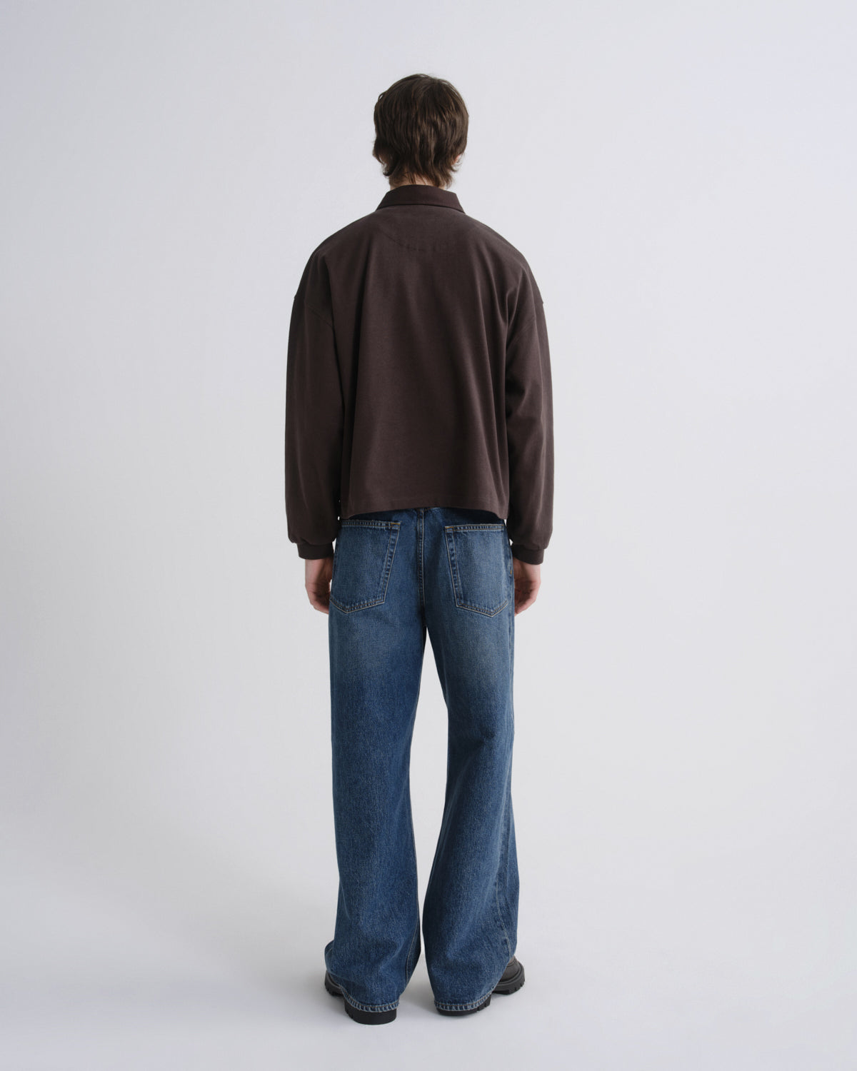 A model shown from the back wearing a dark brown long-sleeve quarter-zip fleece and blue wide-leg jeans, standing against a light gray studio background.