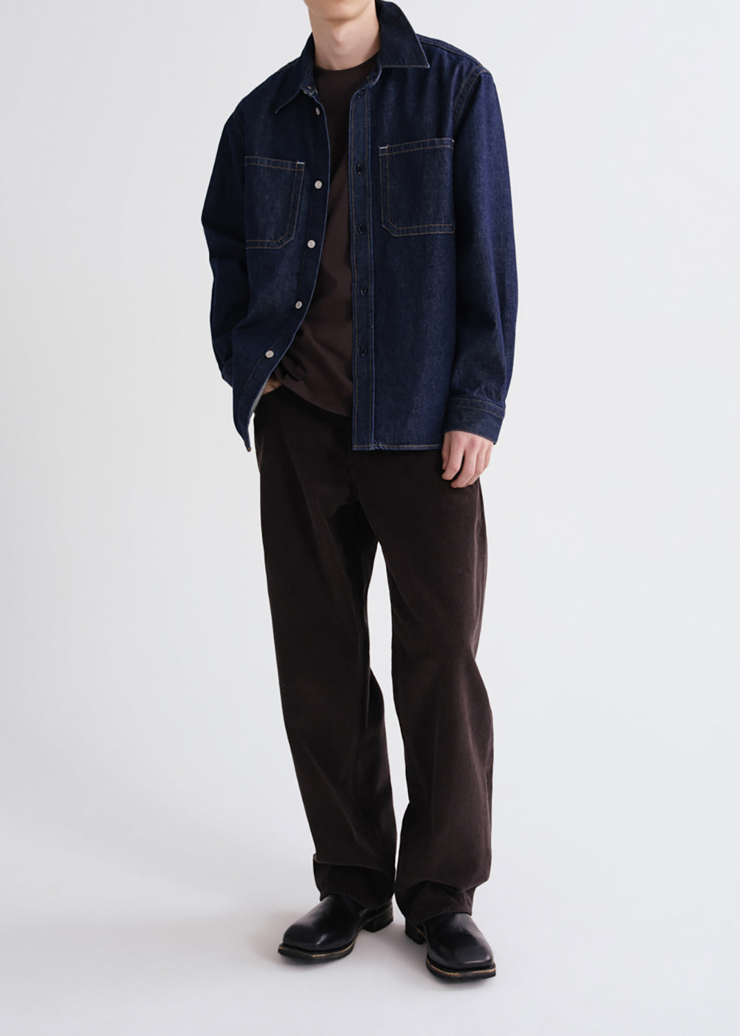 A model wearing a dark indigo denim jacket over a dark brown t-shirt, paired with dark brown corduroy pants and black leather shoes, photographed against a grey background.