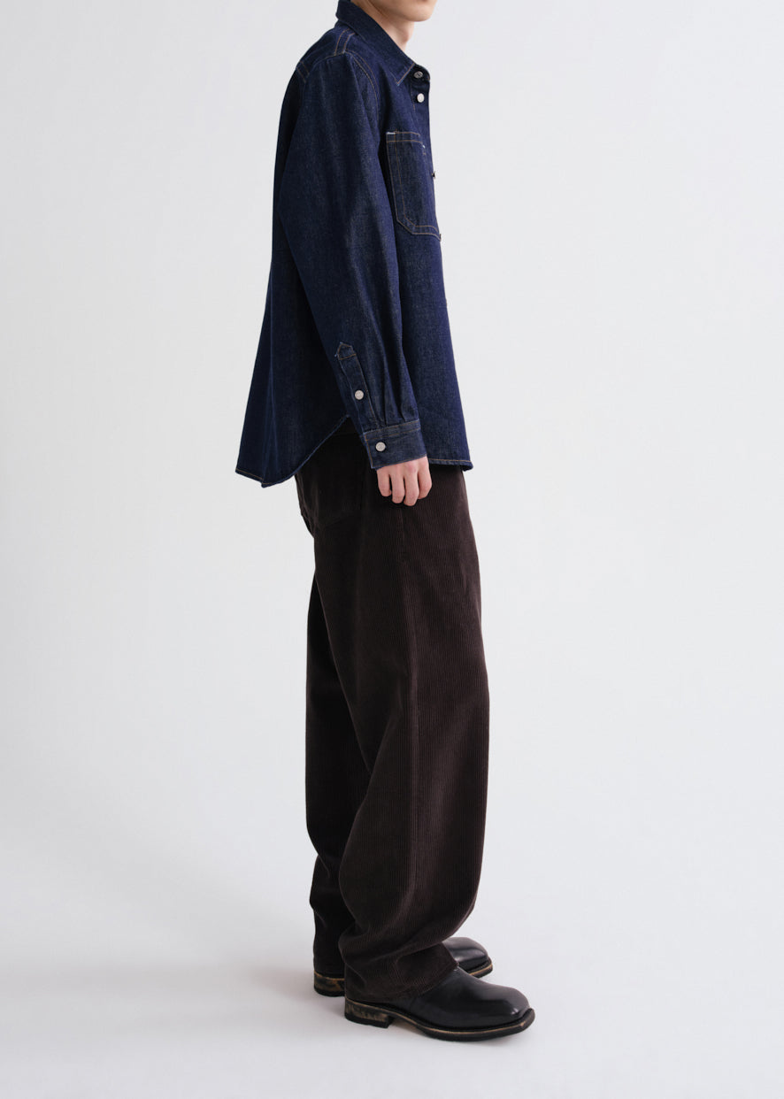 A model shown from the side wearing a dark indigo denim shirt jacket, dark brown corduroy trousers, and black leather boots against a grey background.