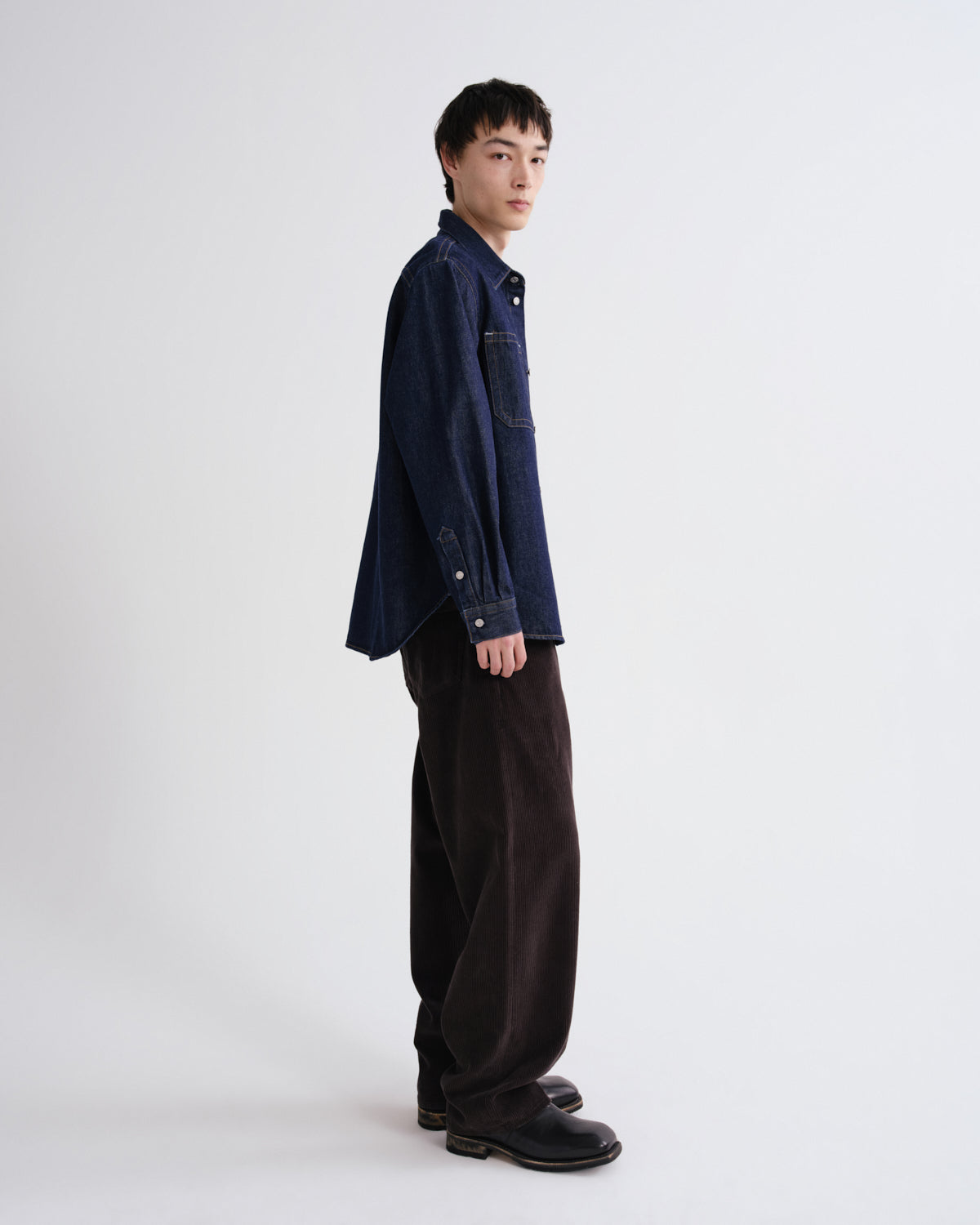 A model stands in profile wearing a dark navy blue denim shirt jacket, loose dark brown corduroy trousers, and black leather shoes against a plain light gray studio background.