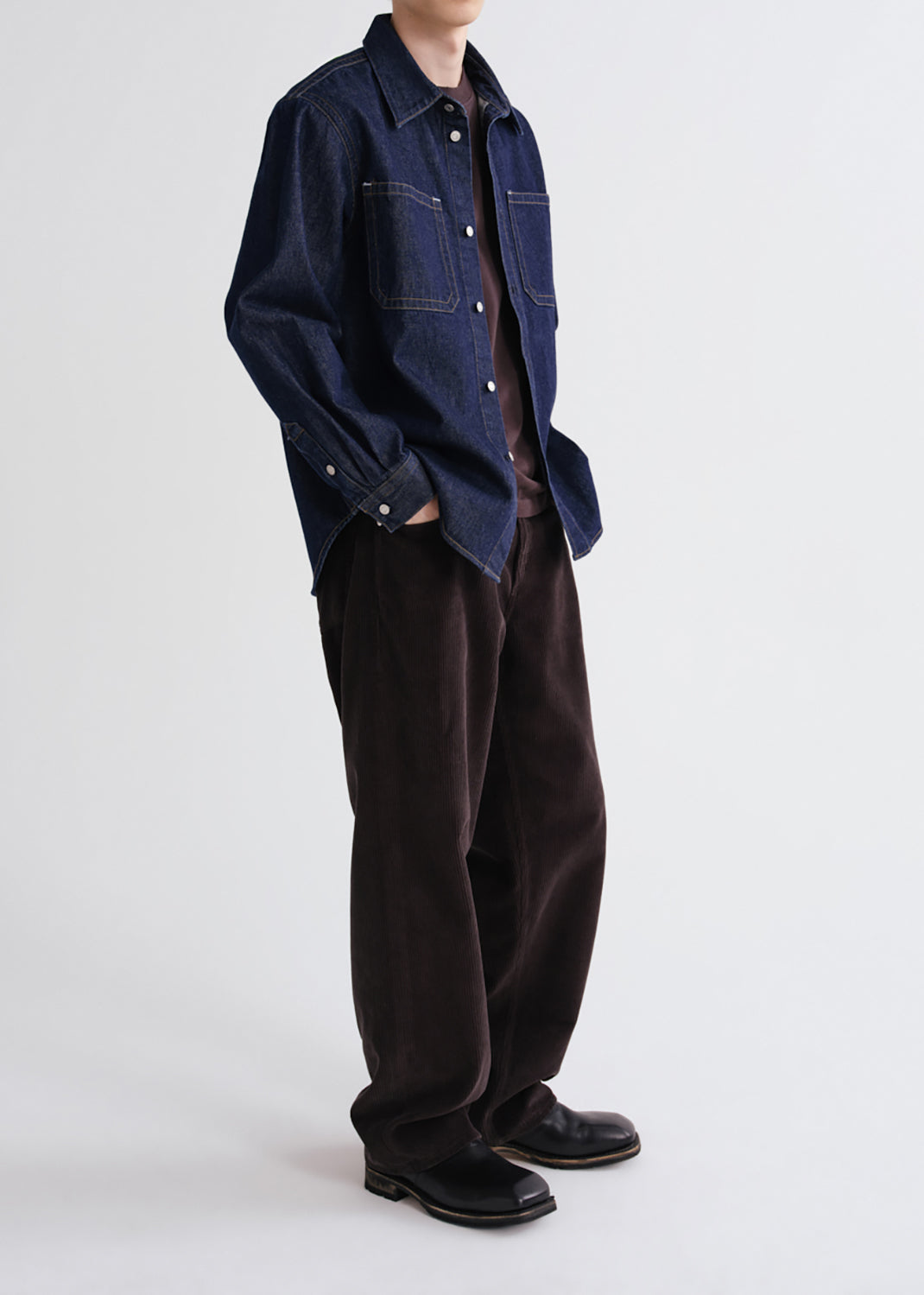 A model wearing a dark blue denim shirt jacket over a brown shirt, paired with dark brown corduroy trousers and black leather shoes, photographed against a grey background.