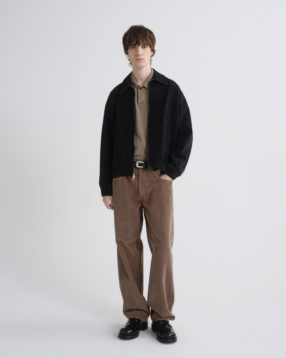 Man wearing a black jacket, brown shirt, and brown pants on a grey background