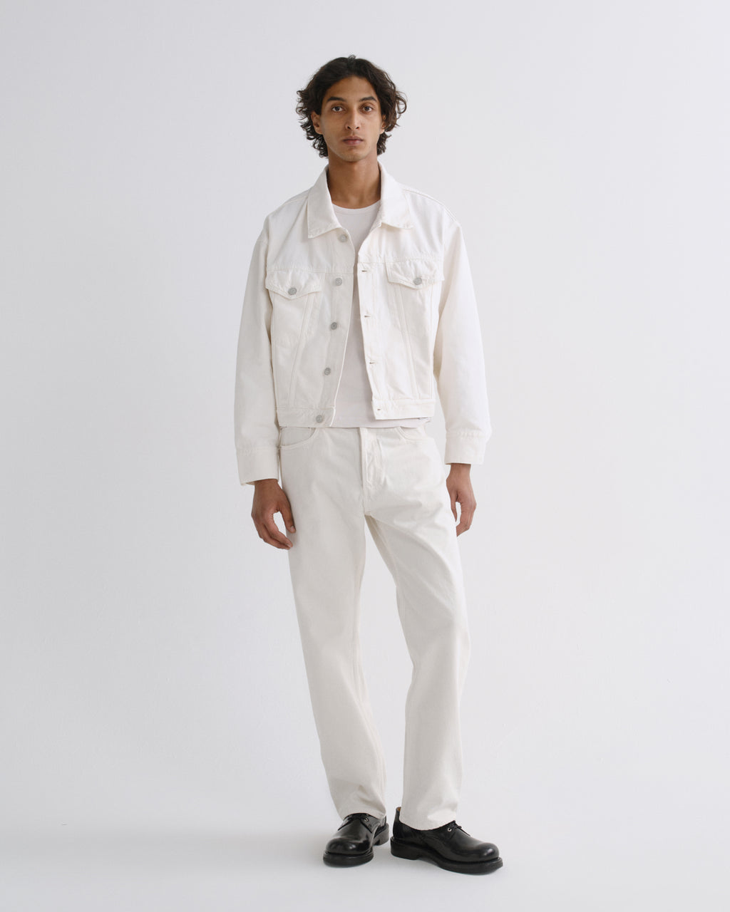 A man modeling a white denim jacket, white shirt, and white trousers with black shoes, standing against a light gray studio background.