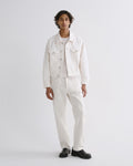 A man modeling a white denim jacket, white shirt, and white trousers with black shoes, standing against a light gray studio background.