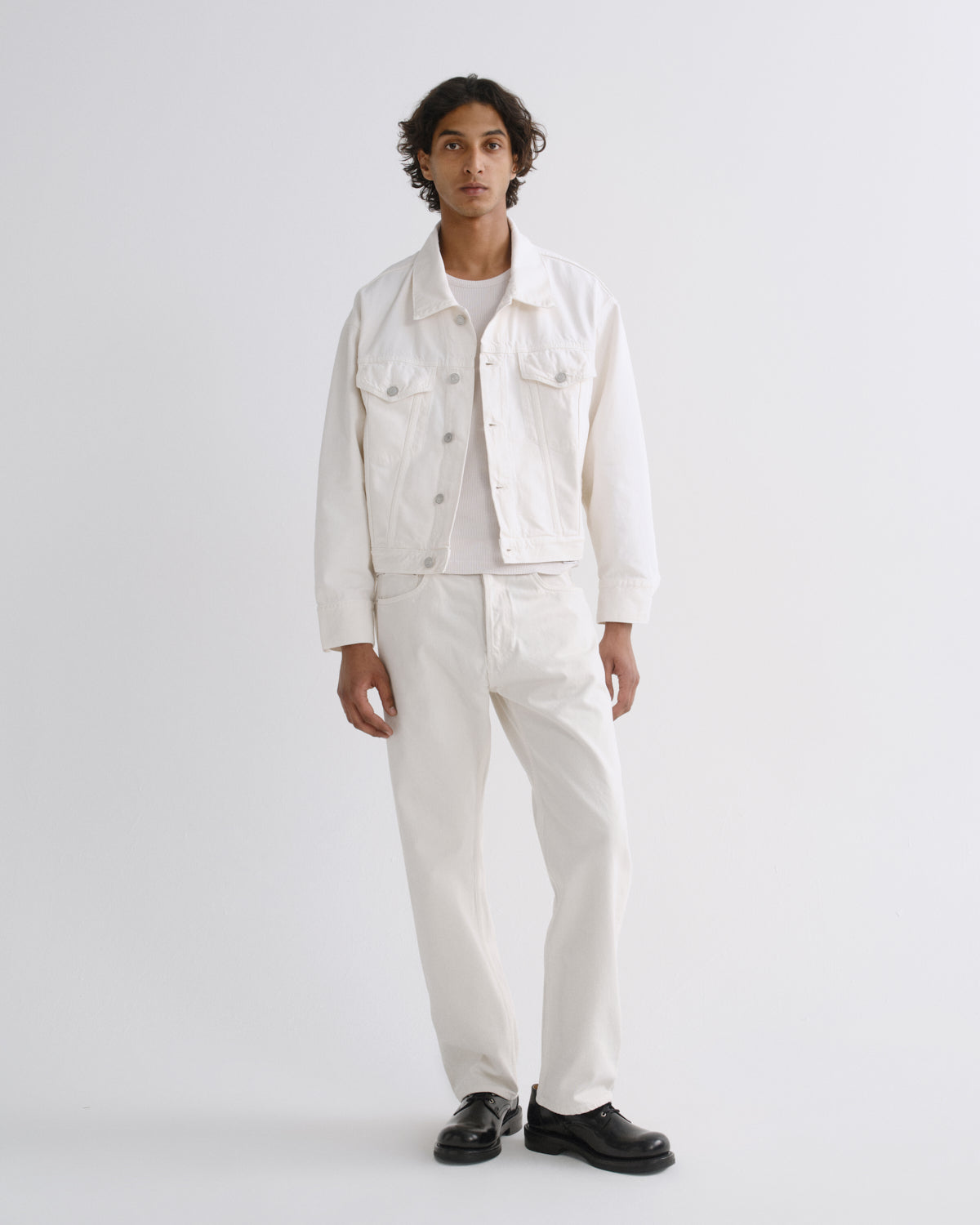 A man modeling a white denim jacket, white shirt, and white trousers with black shoes, standing against a light gray studio background.