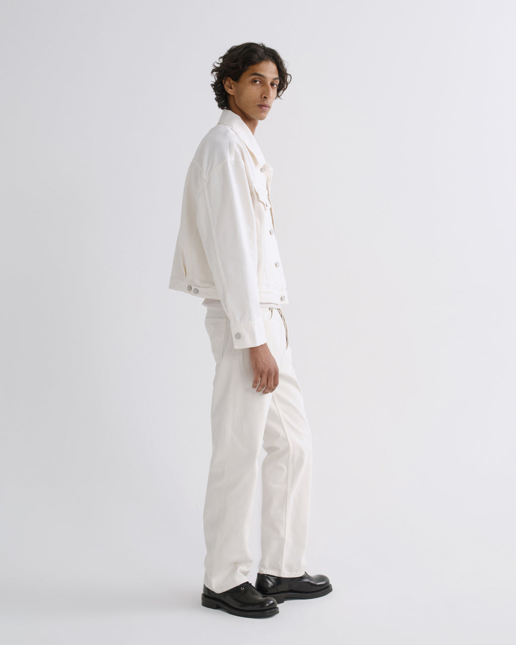 A model wearing a white cropped denim jacket, white wide-leg trousers, and black leather shoes, standing in profile against a light gray studio background.