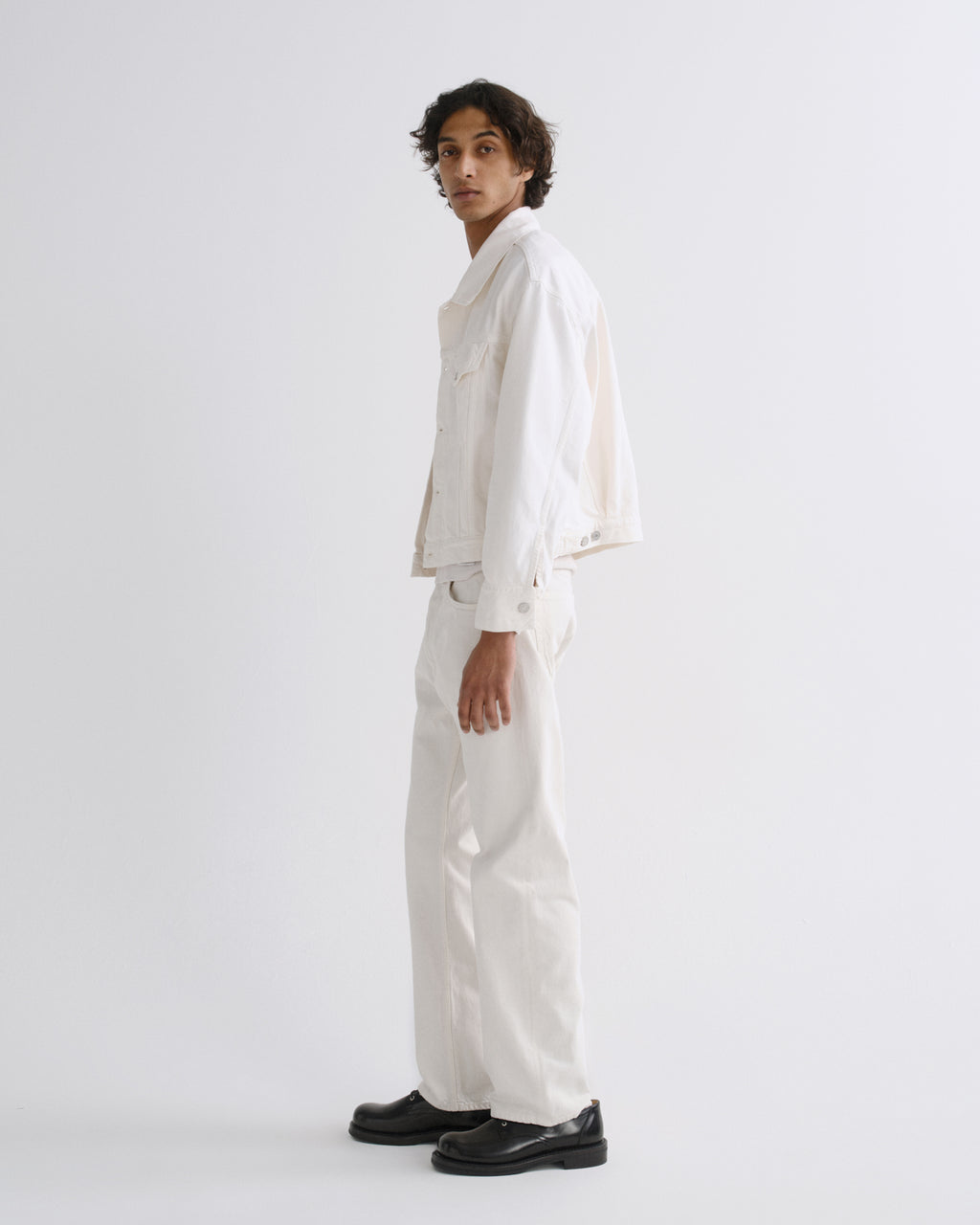 A model wearing a white cropped denim jacket, white wide-leg trousers, and black leather shoes, standing in profile against a light gray studio background.